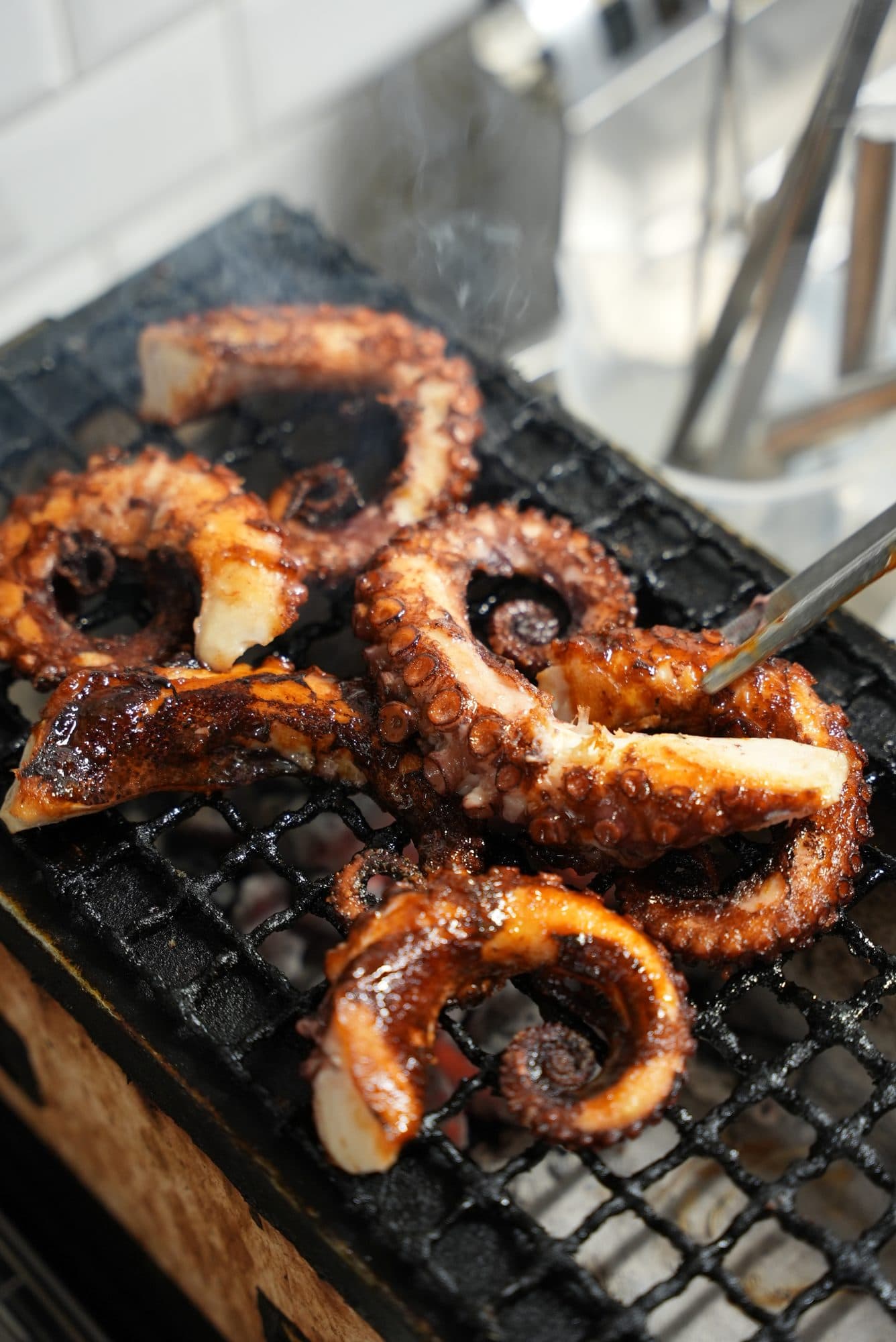 Tentacoli di polpo glassati sulla griglia, sopra la brace accesa, con il fumo che sale e una pinza.