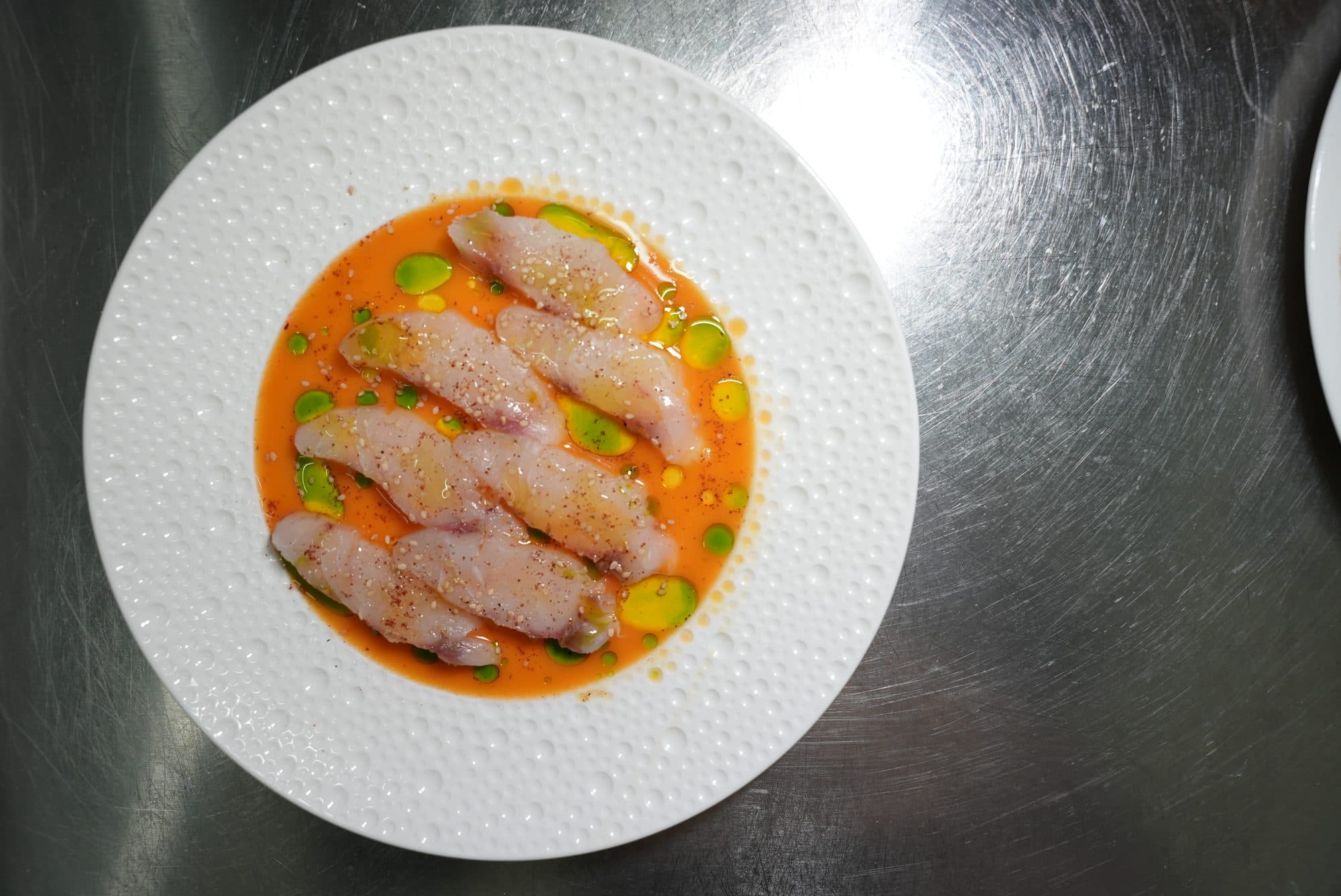 Lo stesso crudo di pesce visto dall’alto, nel piatto bianco su un piano d’acciaio.