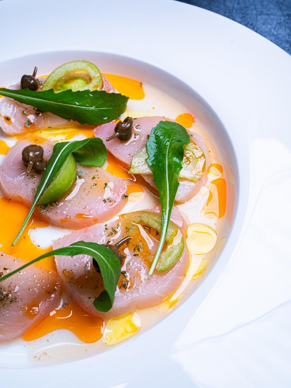Fette di pesce crudo bianco con capperi, rucola, pomodoro verde e un filo d’olio arancione, in un piatto bianco.