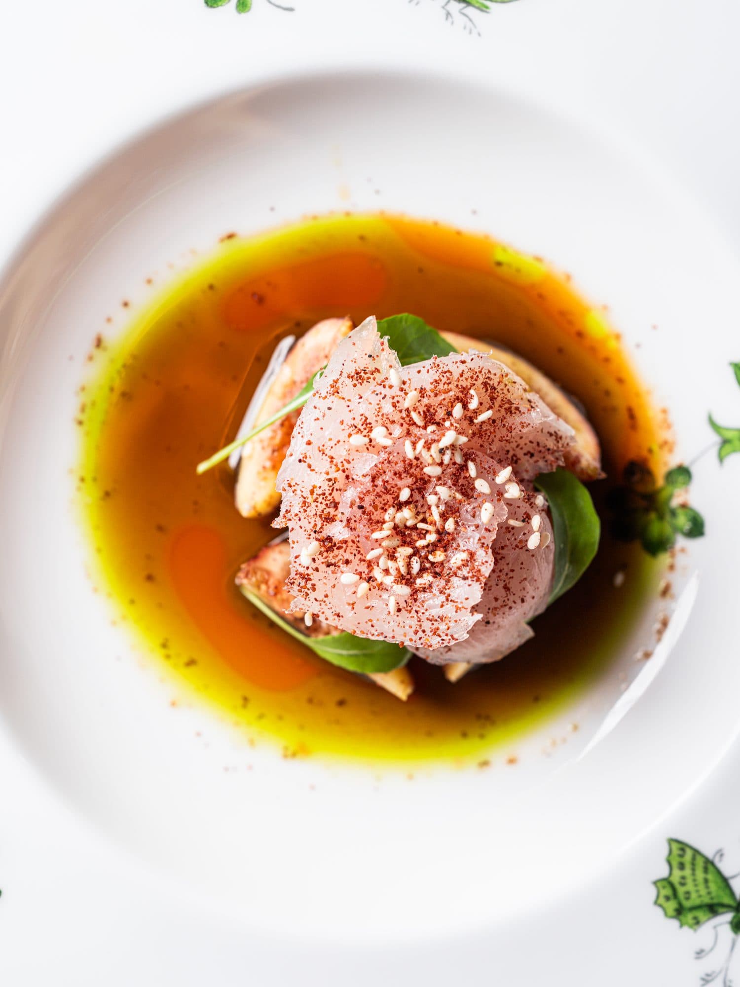 Pesce crudo con semi di sesamo su fette di fico, in un brodo scuro, in un piatto decorato.