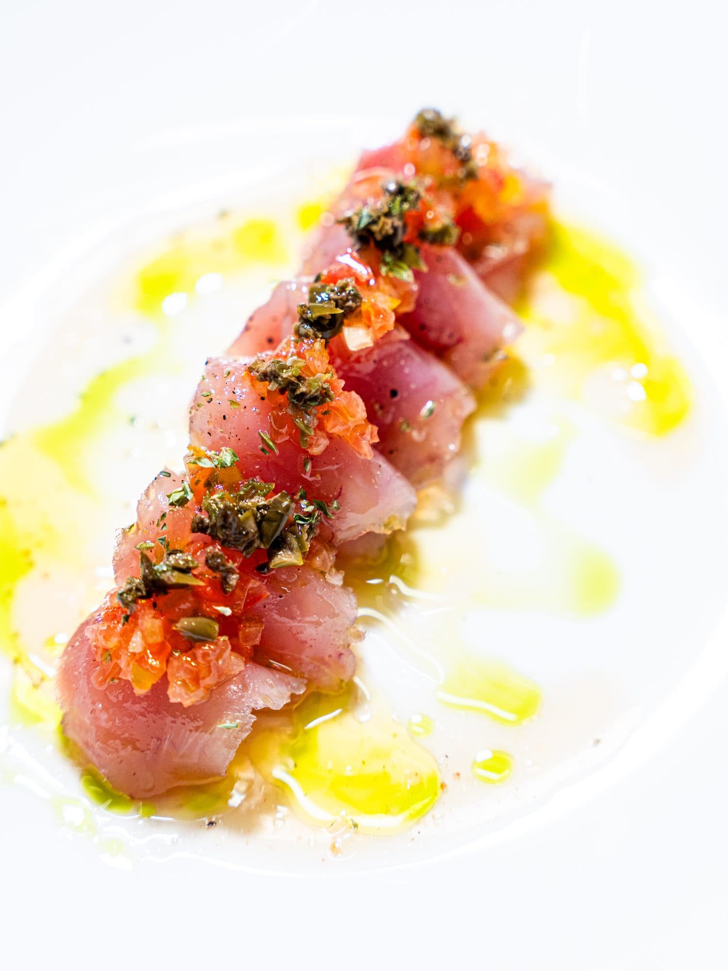 Tonno crudo a cubetti, in fila, con pomodoro, capperi e olio, su un piatto bianco.