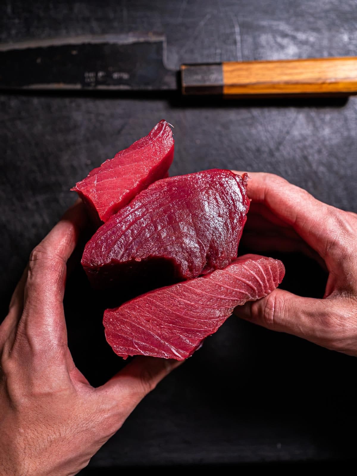 Due mani tengono tre tranci di tonno rosso crudo, sopra un tagliere scuro con un coltello.